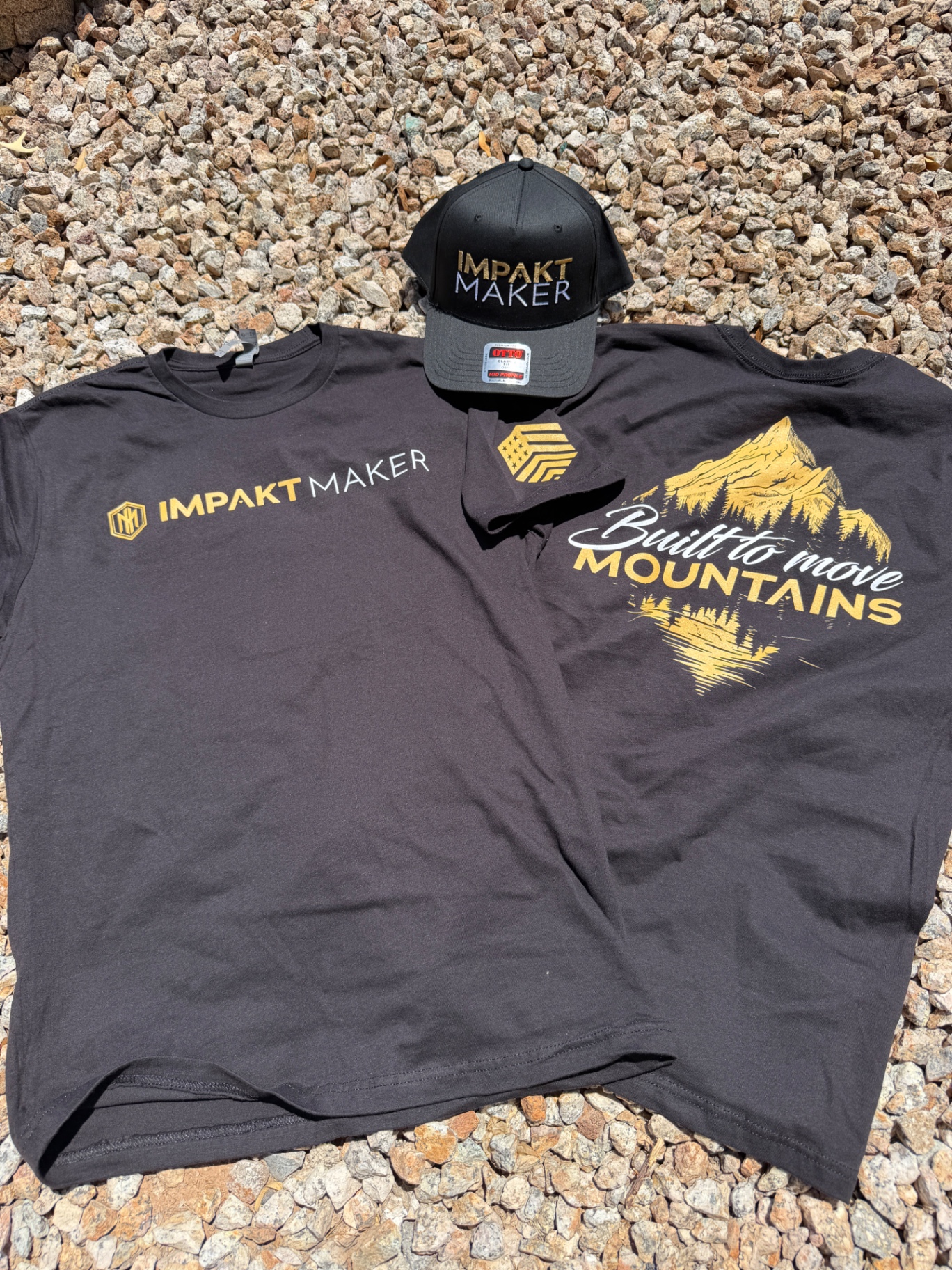 Charcoal Impakt Maker tee with gold and white screen-printed logo and Built to Move Mountains back graphic next to a matching black trucker hat with gold and white embroidered logo, custom apparel set by Colorblox Merch Co. in Gilbert, Arizona